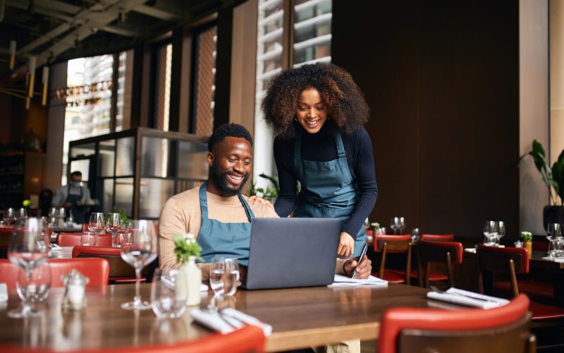 Restaurant owners smiling while reviewing marketing strategies on a laptop inside a bright, modern dining room.
